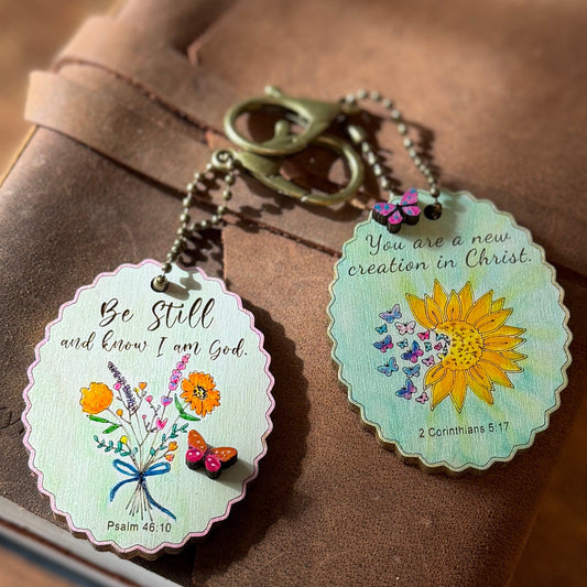 Two decorative keychains with floral designs and inspirational quotes on a brown surface.