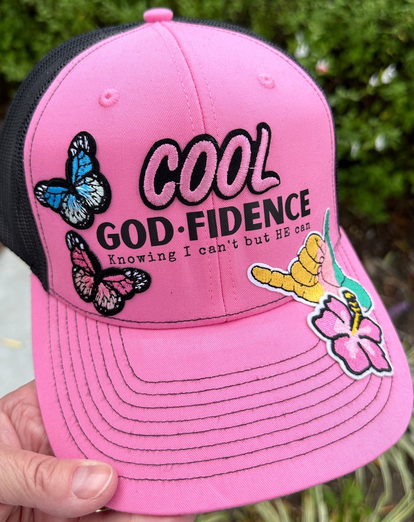 Pink cap with butterfly designs and text held by a hand against a blurred green background