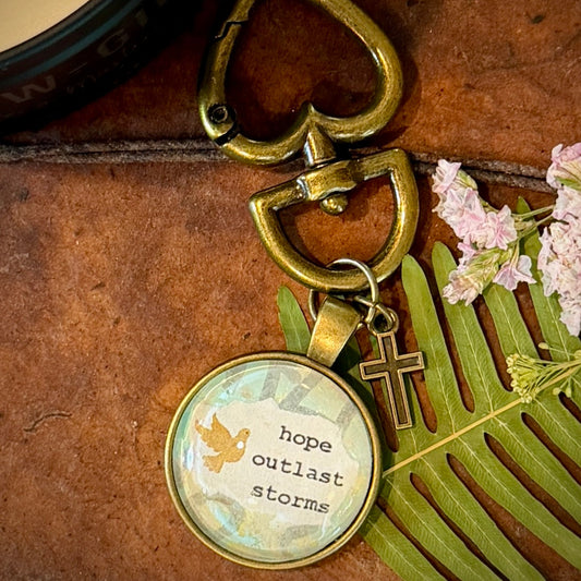 Keychain with 'hope outlast storms' charm on a wooden surface with flowers.