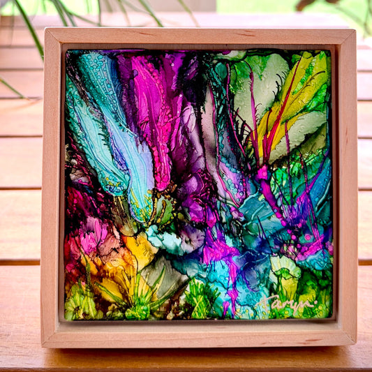 Colorful abstract art piece in a wooden frame on a wooden surface with a blurred natural background