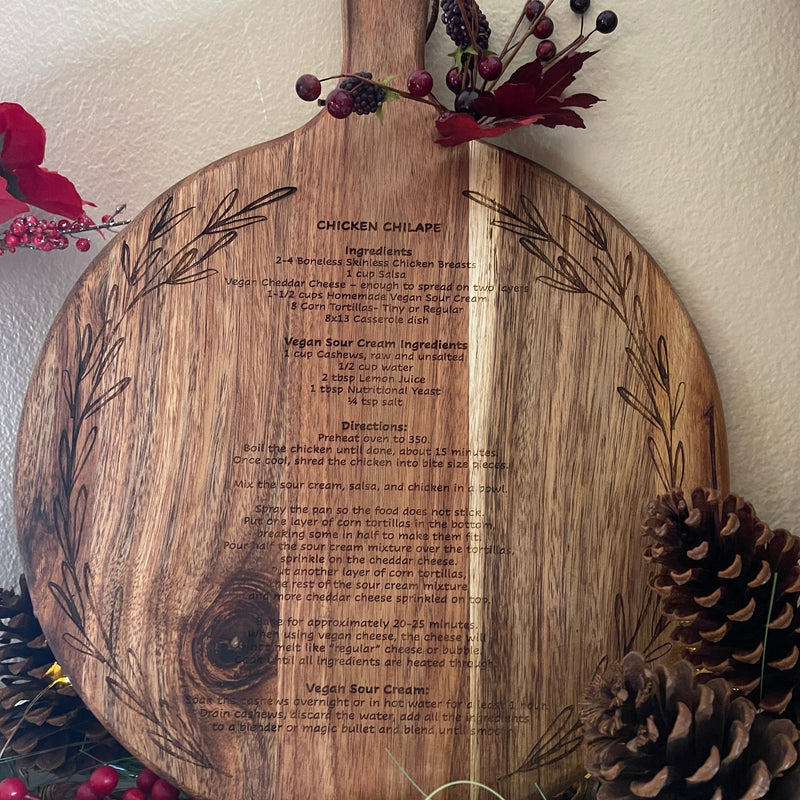 Wooden cutting board with engraved text and floral designs, surrounded by decorative elements like pine cones and berries.