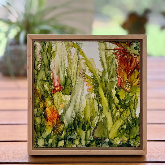 Framed artwork of a nature scene with greenery and a butterfly on a wooden surface.