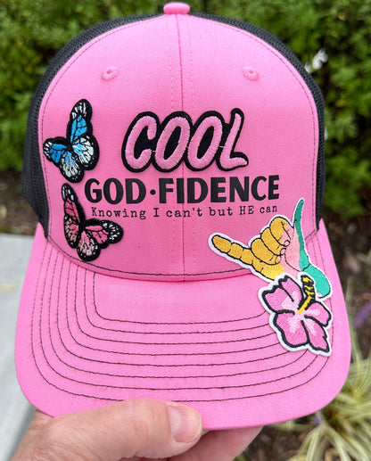 Pink cap with 'Cool God Fidence' text and butterfly designs, held against a blurred green background.