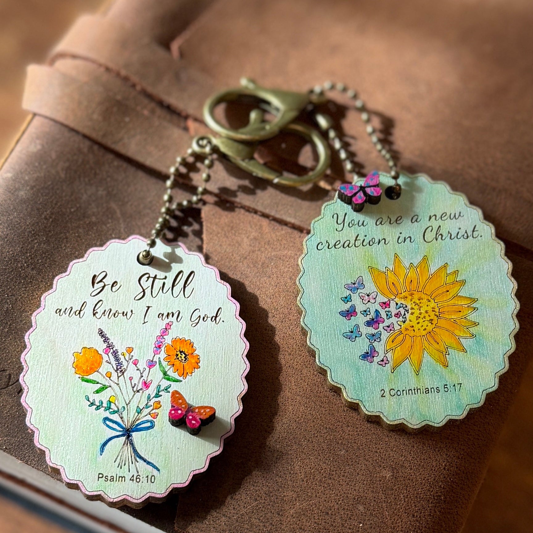 Two decorative keychains with floral designs and inspirational quotes on a brown surface.