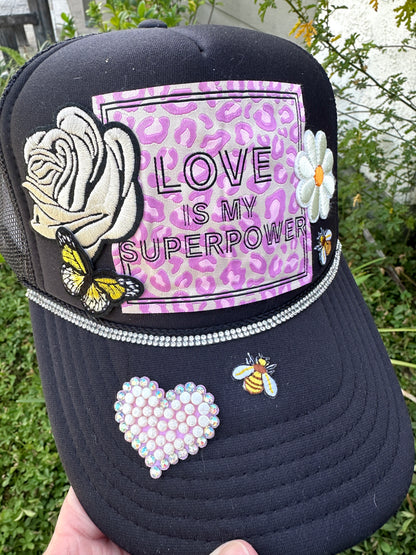 Black cap with decorative patches including a rose, butterfly, 'Love is my Superpower' design, heart, and bee on a blurred green background.