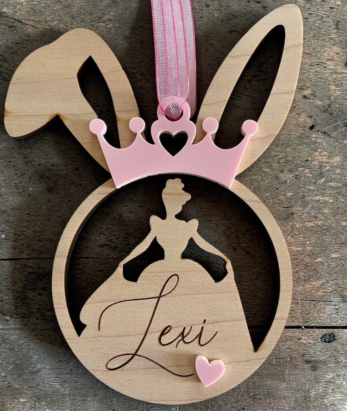Wooden ornament with bunny ears, pink ribbon, and silhouette of a princess dress with 'Lexi' on a wooden surface.