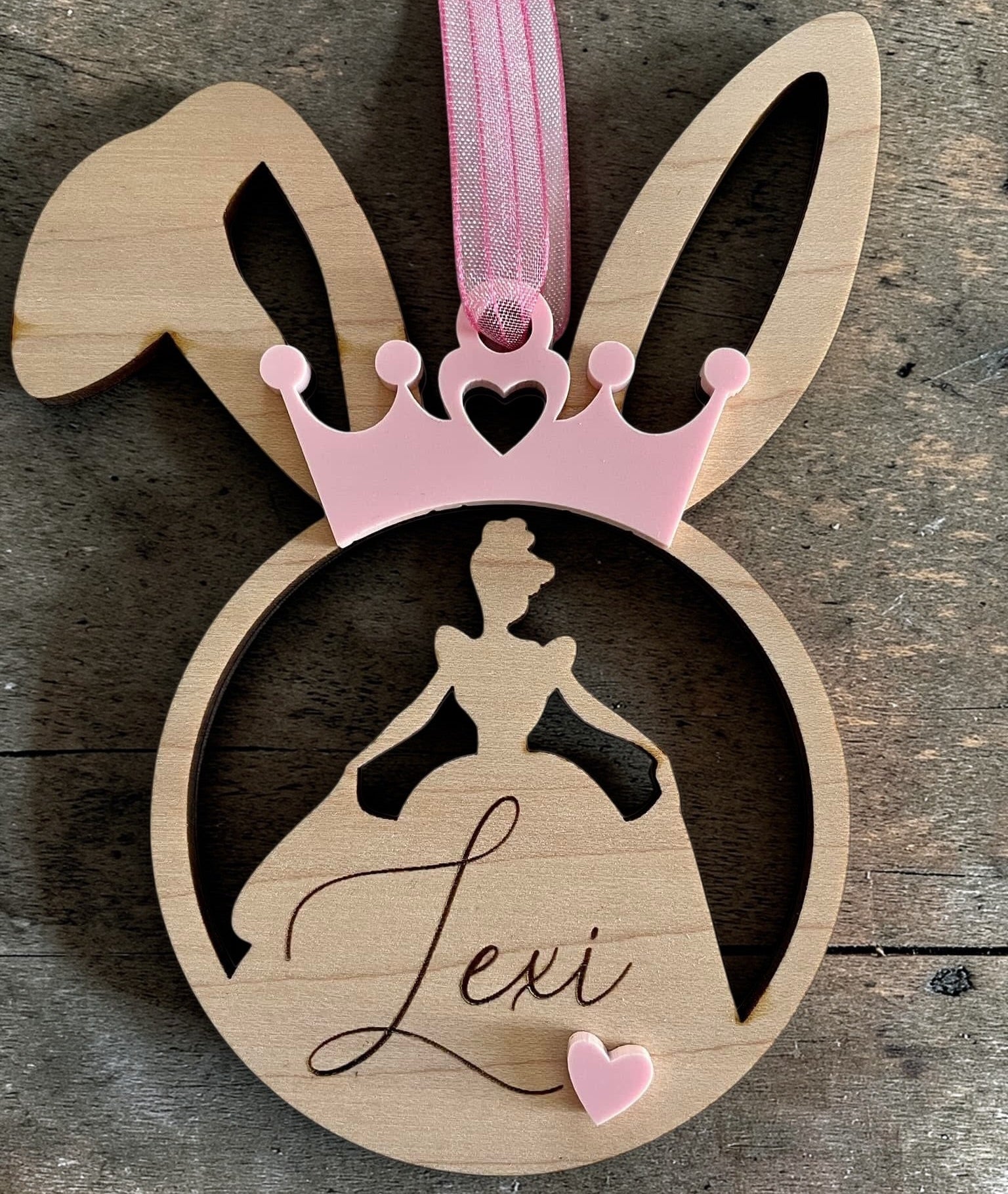 Wooden ornament with bunny ears, pink ribbon, and silhouette of a princess dress with 'Lexi' on a wooden surface.