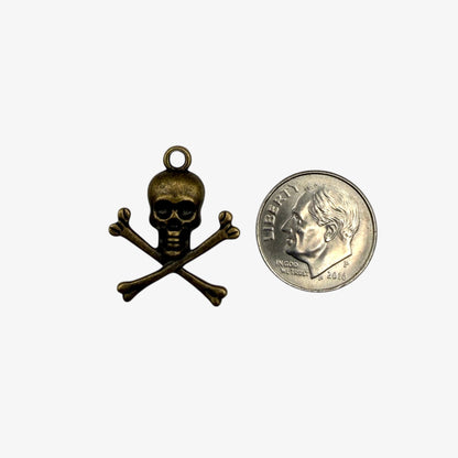 Small brass skull and crossbones pendant next to a U.S. quarter for scale on a white background