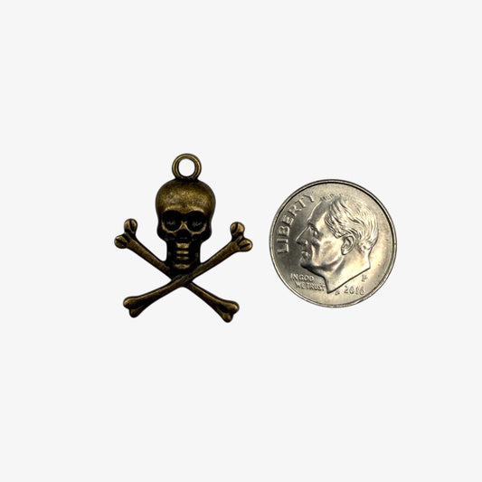 Small brass skull and crossbones pendant next to a U.S. quarter for scale on a white background