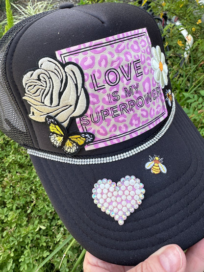 Black cap with decorative elements including a rose, butterfly, and 'Love is my Superpower' text on a purple background.