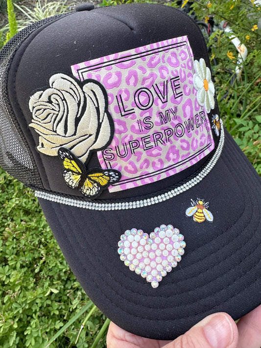 Black cap with decorative elements including a rose, butterfly, and 'Love is my Superpower' text on a purple background.